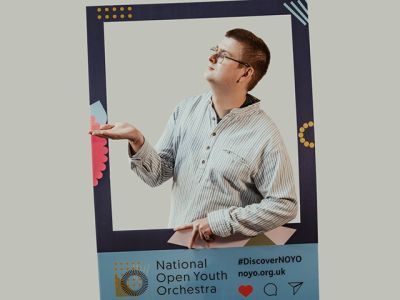 A boy with short brown hair and wearing a white striped shirt stands in between a customised National Open Youth Orchestra Instagram frame. He is looking to his right and has his right hand up and left hand down.